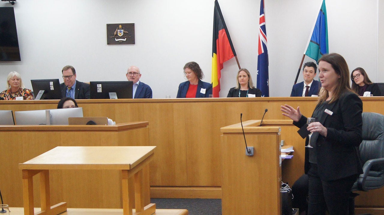 A woman stands in a courtroom at an event, mid-speech. In the background of the image are seven people sitting up on the bench.