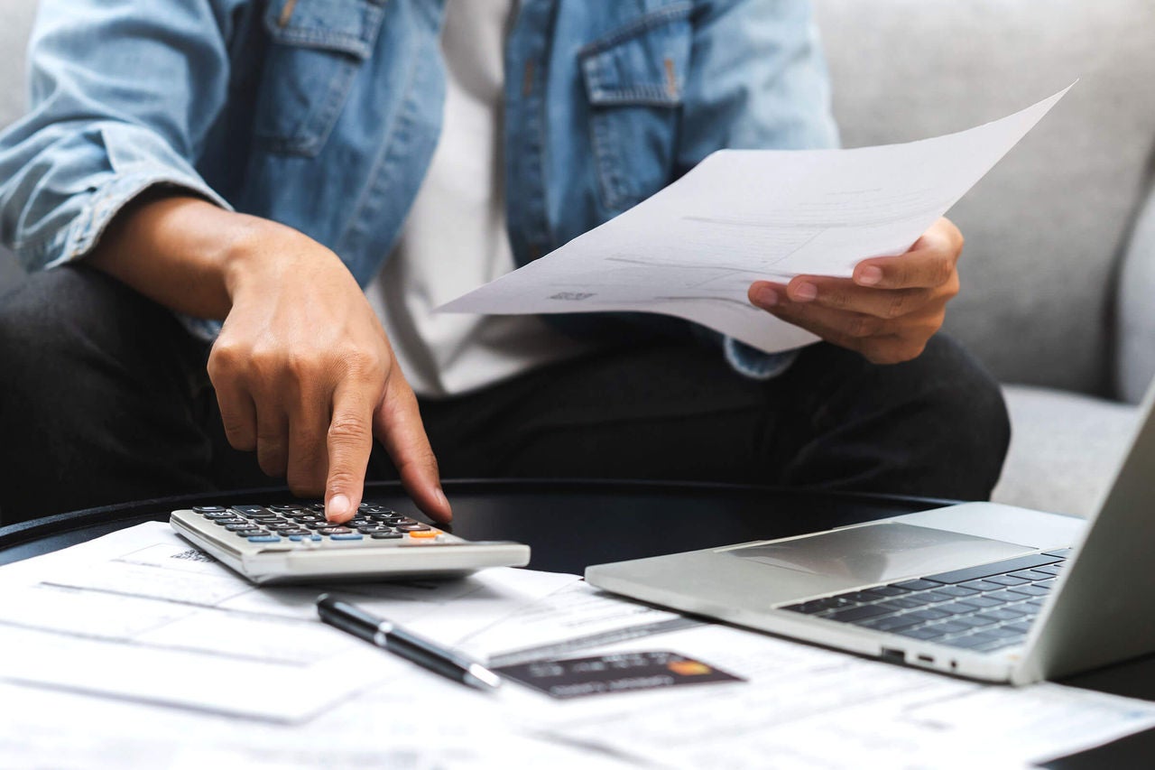 Person sitting on a couch in front of an open laptop on a coffee table, with an unpaid bill in their left hand while their right hand uses a calculator. Additional unpaid bills and a credit card can also be seen on the coffee table.