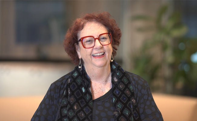 Bev Lazarou smiles to camera. She has red hair and large red glasses.