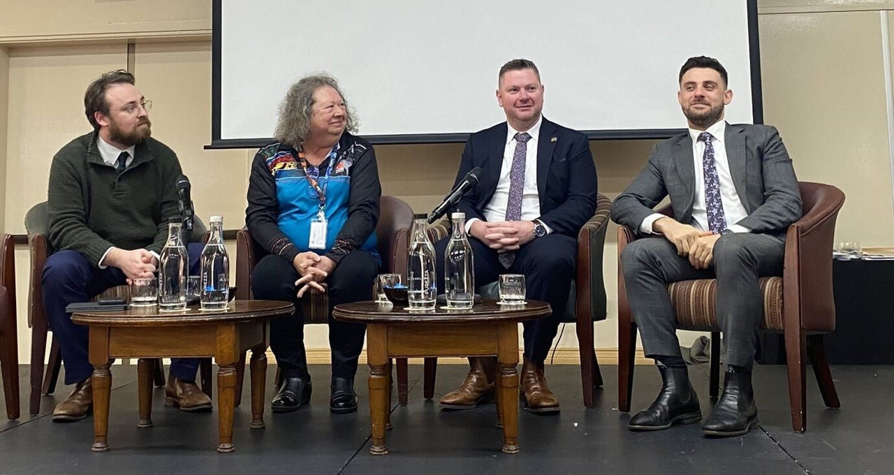 Sam, Anita, Matt and Jordan sit on a stage behind two small wooden tables. Three of the speakers are turned slightly to Jordan. They are all smiling.