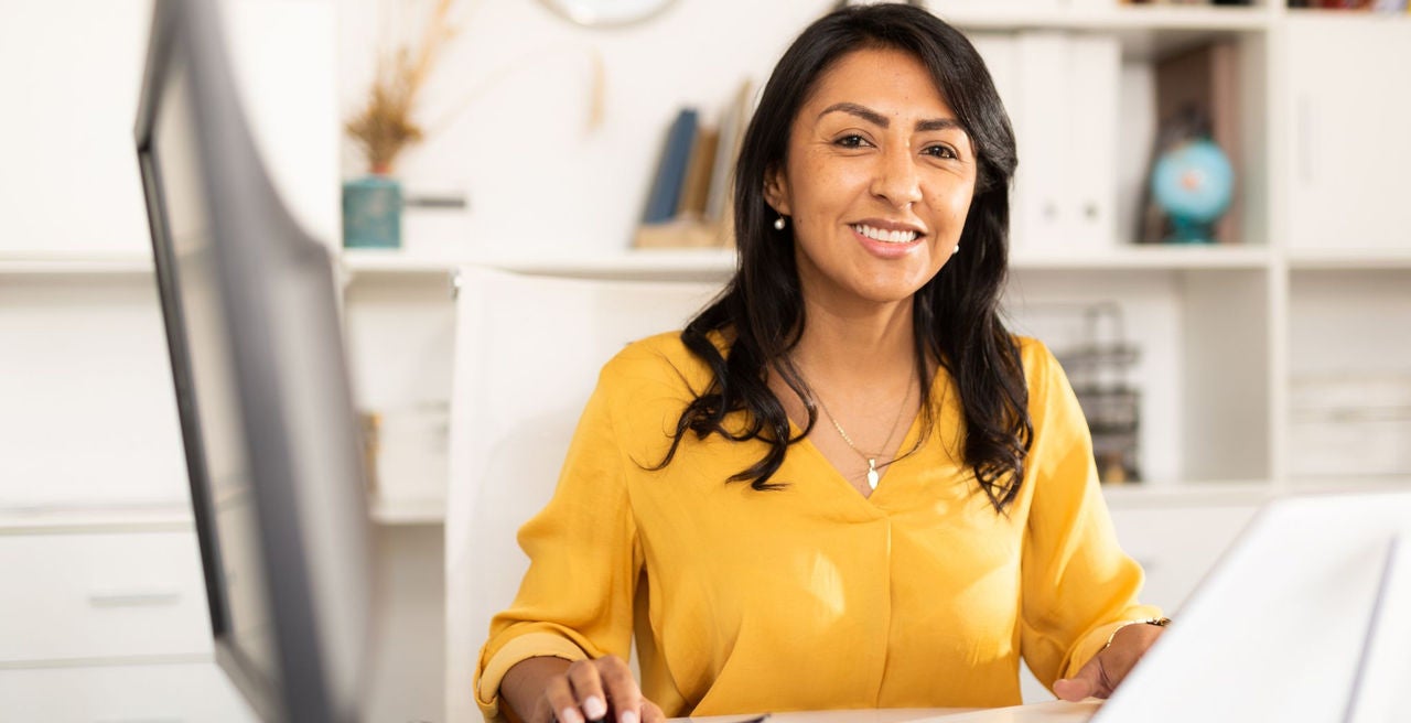 A woman sits at a desk working on a computer. She is wearing a bright yellow shirt and smiling.